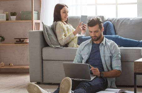 Chica estirada en el sofá mirando el móvil, chico sento en el suelo mirando un portátil